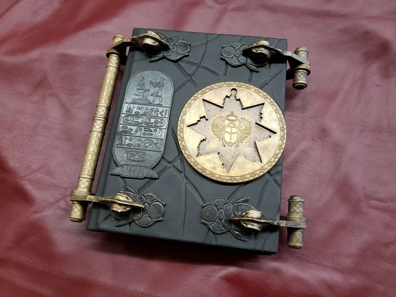 May include: A black leather-bound journal with gold metal clasps and a decorative star-shaped design with an Egyptian scarab beetle motif. The journal has a textured surface and a raised design.