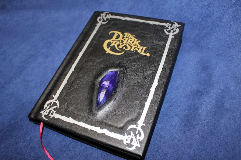 May include: A black leather-bound journal with a silver border and a raised, purple crystal on the front cover. The title "The Dark Crystal" is embossed in gold on the cover.