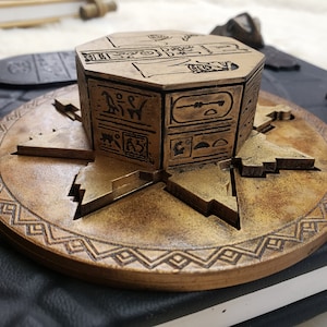 May include: A wooden, octagonal puzzle box with intricate carvings of Egyptian hieroglyphs. The box is sitting on a round, wooden base with a decorative border.