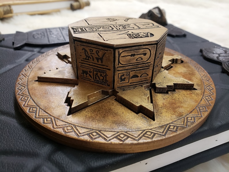 May include: A wooden, octagonal puzzle box with intricate carvings of Egyptian hieroglyphs. The box is sitting on a round, wooden base with a decorative border.