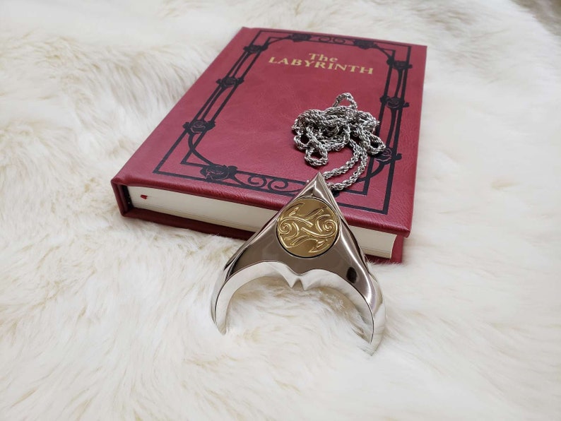 May include: A silver necklace with a gold pendant shaped like a crescent moon with a swirling design. The necklace is lying on a white fluffy surface next to a red book with the title "The Labyrinth" on the cover.