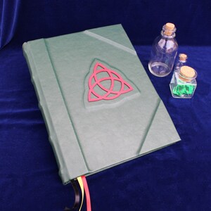 May include: A green leather-bound journal with a red triquetra symbol on the cover. The journal has three ribbon bookmarks in black, yellow, and red.