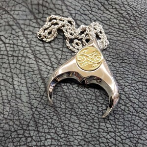 May include: A silver pendant necklace with a gold-toned, circular design in the center. The pendant is shaped like a stylized manta ray with two curved, pointed fins. The necklace is on a silver chain.