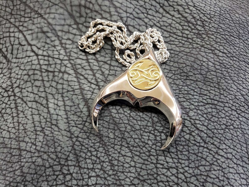 May include: A silver pendant necklace with a gold-toned, circular design in the center. The pendant is shaped like a stylized manta ray with two curved, pointed fins. The necklace is on a silver chain.