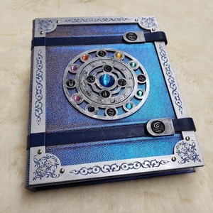 May include: A blue leather-bound journal with a silver metal design featuring a circular compass with colored gems. The journal has a decorative silver border and a blue ribbon closure.