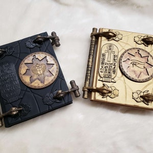 May include: Two decorative journals with Egyptian-themed designs. One journal is black with gold accents and features a scarab beetle. The other journal is gold with black accents and features an eye of Horus.