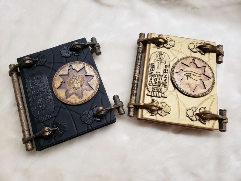 May include: Two decorative journals with Egyptian-themed designs. One journal is black with gold accents and features a scarab beetle. The other journal is gold with black accents and features an eye of Horus.