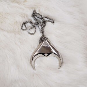 May include: A silver necklace with a large, abstract pendant shaped like a crescent moon. The pendant is made of smooth, polished metal and has a slightly textured surface.