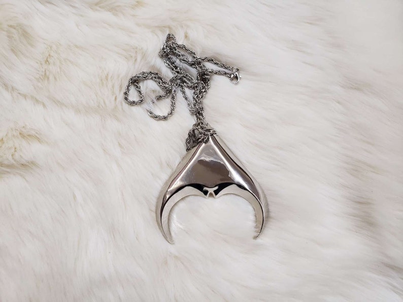 May include: A silver necklace with a large, abstract pendant shaped like a crescent moon. The pendant is made of smooth, polished metal and has a slightly textured surface.