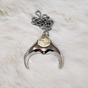 May include: A silver pendant necklace with a gold circular design in the center. The pendant is shaped like a crescent moon with a pointed tip.