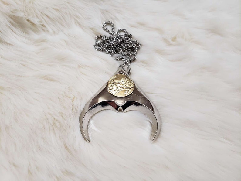 May include: A silver pendant necklace with a gold circular design in the center. The pendant is shaped like a crescent moon with a pointed tip.