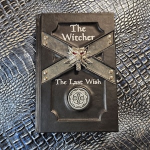 Puede incluir: Un libro negro titulado The Witcher The Last Wish con un emblema de cabeza de lobo plateado con ojos rojos. La cubierta tiene dos correas metálicas cruzadas con remaches y un medallón circular plateado con un diseño geométrico intrincado.