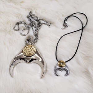 May include: Two silver necklaces with crescent moon pendants. The pendants have a gold-colored design in the center.