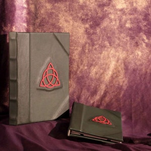 May include: Two green leather-bound journals with a red triquetra symbol on the front cover. The journals are on a purple background.
