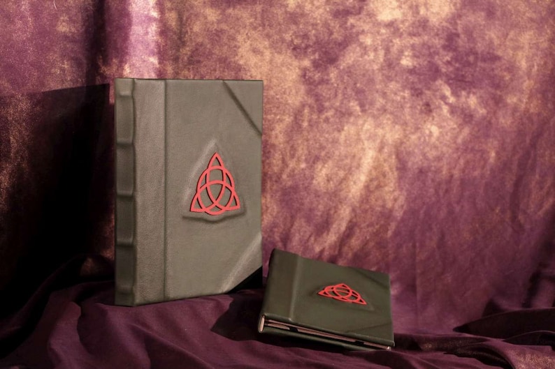 May include: Two green leather-bound journals with a red triquetra symbol on the front cover. The journals are on a purple background.