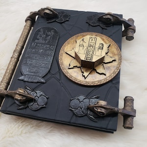 May include: A black leather-bound journal with gold accents and intricate details. The journal has a unique design with a circular, gold-colored, Egyptian-themed lock and key. The journal has two gold-colored metal clasps on each side.