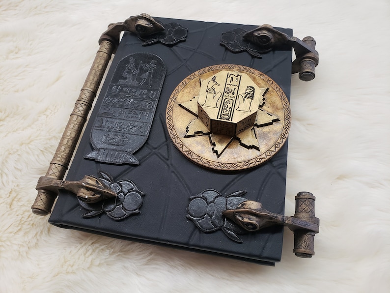 May include: A black leather-bound journal with gold accents and intricate details. The journal has a unique design with a circular, gold-colored, Egyptian-themed lock and key. The journal has two gold-colored metal clasps on each side.