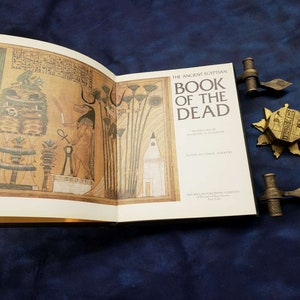 May include: An open book titled "The Ancient Egyptian Book of the Dead" with gold-colored edges and a black cover. The book is open to a page with an illustration of an Egyptian god and hieroglyphics. The book is resting on a blue surface with two gold-colored bookends.
