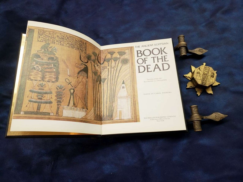 May include: An open book titled "The Ancient Egyptian Book of the Dead" with gold-colored edges and a black cover. The book is open to a page with an illustration of an Egyptian god and hieroglyphics. The book is resting on a blue surface with two gold-colored bookends.