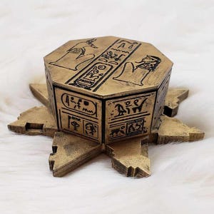 May include: A gold-coloured, octagonal box with intricate Egyptian hieroglyphics and figures. The box sits on a decorative, star-shaped base.