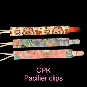 Puede incluir: Tres clips de chupete CPK con diferentes patrones. Uno tiene un fondo blanco con fresas y una niña con gorro. Otro tiene un fondo azul con osos de dibujos animados. El último tiene un fondo rosa con osos de dibujos animados.