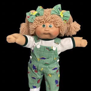 May include: A doll with tan yarn hair styled in pigtails with green striped bows. The doll has green eyes, a round face, and wears a white shirt under green striped overalls featuring a colourful dinosaur print and white shoes.
