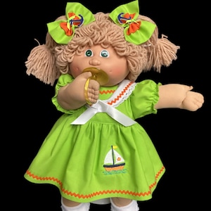 Pode incluir: Boneca Cabbage Patch Kid vintage vestida com um vestido verde limão com um desenho de veleiro. A boneca tem cabelo loiro de lã, olhos verdes e uma chupeta. O vestido tem acabamentos laranja e uma gola branca com renda.