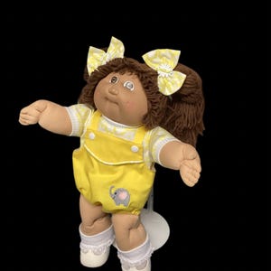 May include: A vintage doll with brown hair in pigtails, decorated with yellow and white patterned bows. The doll has brown skin and wears a yellow romper with an elephant applique. White socks and shoes complete the outfit.