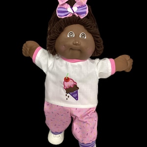 Puede incluir: Una muñeca con cabello de lana marrón oscuro peinado en una coleta alta con dos lazos a rayas rosas y morados con corazones blancos. La muñeca viste una camiseta blanca de manga corta con un gráfico de helado y pantalones con estampado de chispas.