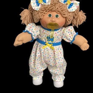 May include: A vintage Cabbage Patch Kids doll wearing a white onesie with colorful polka dots, blue trim, and a yellow bow. The doll has blonde yarn hair, green eyes, and two white bows with blue polka dots. It has a pacifier in its mouth.