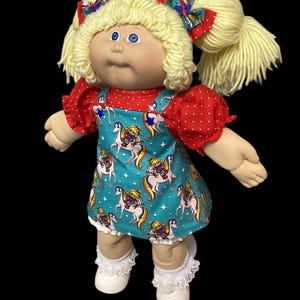 May include: A doll with blonde yarn hair styled in pigtails with red and patterned bows. It wears a red polka-dot shirt under a teal jumper featuring a repeating print of figures riding unicorns. The doll has white shoes with lace-trimmed socks.