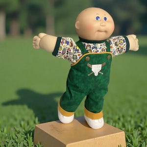May include: A bald doll with blue eyes and rosy cheeks wearing a white long-sleeve shirt with a colorful sports-themed pattern and dark green overalls. The overalls feature a baseball home plate and bat graphic on the chest with gold buttons.