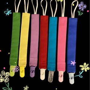 以下が含まれることがあります： 緑、黄色、紫、赤、ターコイズ、ピンク、青の7色のカラフルなおしゃぶりクリップのコレクション。各クリップには、お揃いの生地ストラップ、白いループ、プラスチッククリップが付いています。背景は、描かれた花のある黒色です。