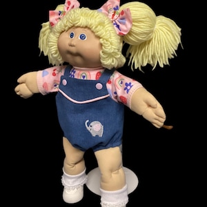 May include: A vintage Cabbage Patch Kid doll with blonde yarn hair styled in pigtails, adorned with pink and blue patterned bows. The doll wears a pink shirt with a rainbow and fruit pattern, denim overalls with an embroidered elephant, white socks, and shoes.