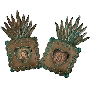 May include: Two square metal wall plaques featuring a bronze finish with teal accents and a flame-like top. Each plaque has a central heart-shaped cutout containing a small image of the Virgin Mary, surrounded by decorative scrollwork edges.