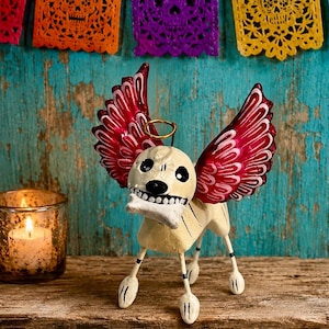 May include: A cream-colored metal dog figurine with a skeletal face, black eye sockets, and a wide grin holding a white bone. The dog features large, vibrant red wings with white scalloped details and a thin gold wire halo above its head.