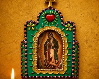 May include: A decorative wall plaque featuring a central image of the Virgin of Guadalupe framed in gold, surrounded by a green border with pink and blue dots, a red heart at the top, and a gold cross finial, set against a textured yellow wall.