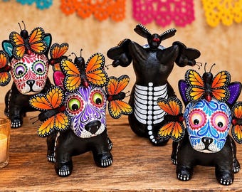 May include: Four black dog figurines with painted faces and orange butterfly accents. Three dogs feature colorful floral patterns on their heads, while one shows a white skeletal ribcage design on its back. Each dog is adorned with monarch butterflies.