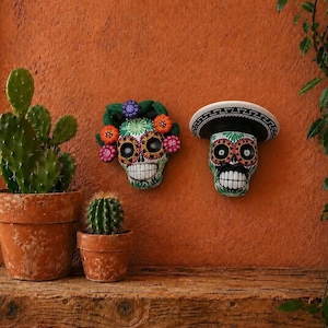May include: Two Day of the Dead skull decorations. One skull has a floral crown, the other wears a sombrero. Both are painted with colorful patterns and sit against an orange wall. A cactus and a small potted cactus are in the foreground.