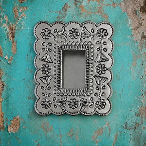 May include: A rectangular silver-toned metal picture frame featuring a scalloped edge and an embossed floral pattern. The frame has a central rectangular opening and a decorative border with repeating flower and leaf motifs in a shiny finish.