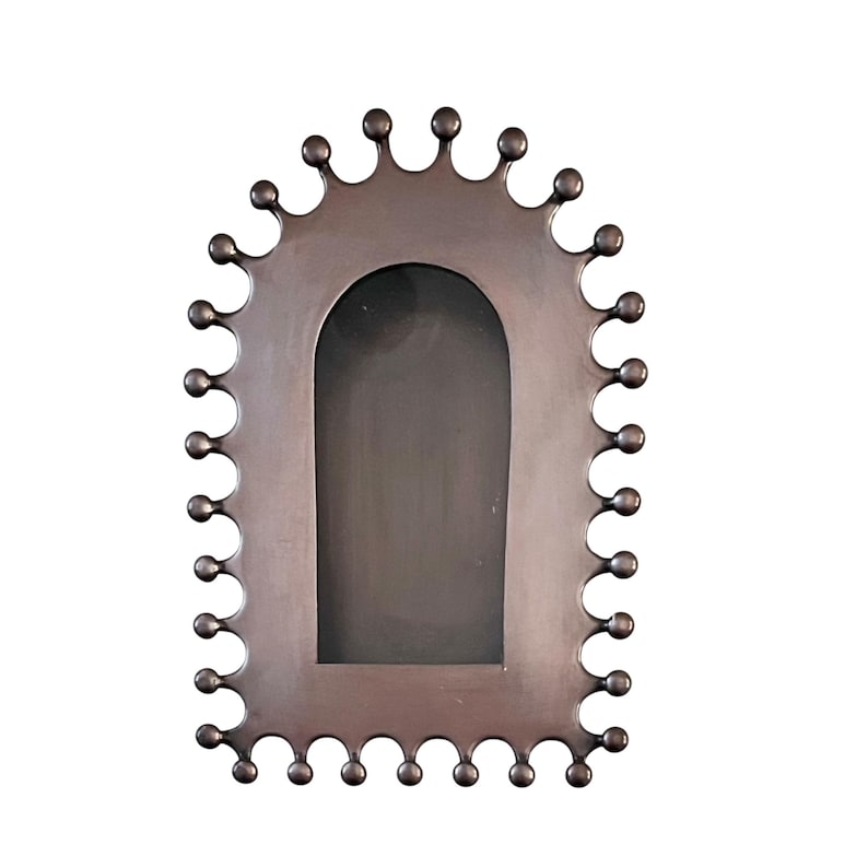 May include: A bronze-colored metal picture frame with a unique scalloped edge design. The frame has a rectangular shape with a rounded top and a dark interior space for a photo. The edge is adorned with small, round, decorative accents.