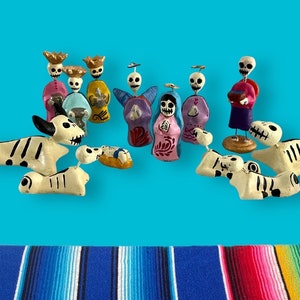 May include: A collection of Day of the Dead figurines, including skeleton figures and dogs, in various colors. The figurines are set against a bright blue background, with a colorful striped textile at the bottom.