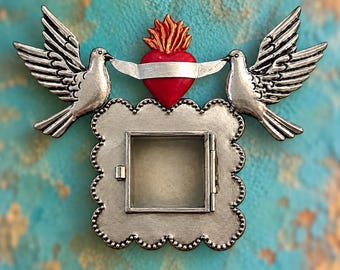 May include: A silver-toned metal wall hanging featuring a square, hinged frame with a scalloped edge. Above the frame, two silver doves hold a red heart with a gold flame and a silver banner in their beaks, creating a symmetrical design.