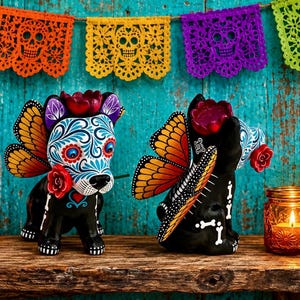 May include: Two black dog figurines decorated in a Day of the Dead style with intricate blue and white floral patterns on their faces. Each dog features orange and black monarch butterfly wings, red flower accents, and white bone motifs on their bodies.