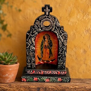 May include: A black arched retablo featuring a central painting of the Virgin of Guadalupe. The frame is decorated with silver-toned metal milagros and a cross at the top. The base is painted with red and green floral patterns.