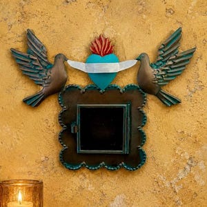 May include: A decorative wall hanging featuring two bronze-colored birds with teal-tinted wings flanking a central turquoise heart topped with a red flame. Below the heart is a square, dark-toned metal frame with a small hinged door in the center.