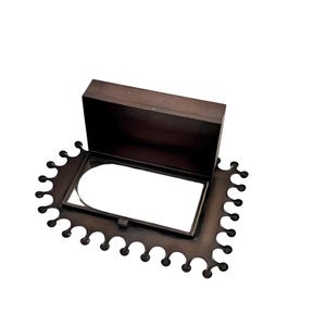 May include: A dark brown, rectangular box with a hinged lid, set on a decorative, scalloped-edge base. The box is open, revealing a white interior. The base and box appear to be made of metal.