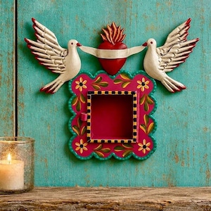 May include: A decorative wall-mounted metal box featuring a vibrant pink square frame with hand-painted yellow flowers and green leaves. Above the frame, two white doves with outstretched wings flank a central red heart topped with orange flames.