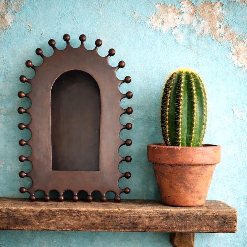May include: A decorative, arched metal frame with a scalloped edge and small ball accents. Beside it, a green cactus with yellow spines sits in a terracotta pot. The items rest on a weathered wooden shelf against a blue wall.