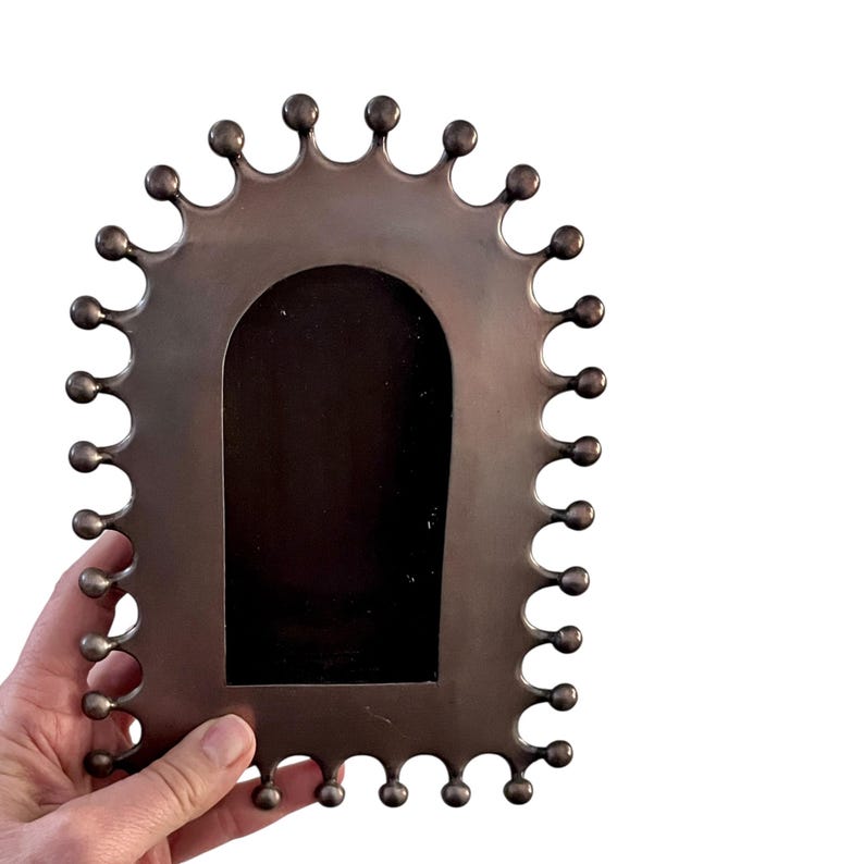 May include: Bronze-colored metal picture frame with a unique scalloped edge design. The frame has a dark interior and is held by a hand against a white background. The frame's edge features a series of rounded, ball-like accents.
